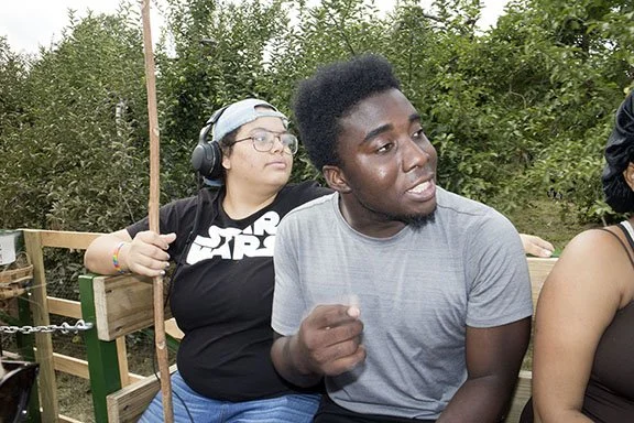 Two people sitting outdoors next to a green hedge, one wearing headphones and a Star Wars shirt, the other wearing a gray t-shirt and gesturing with their hand.