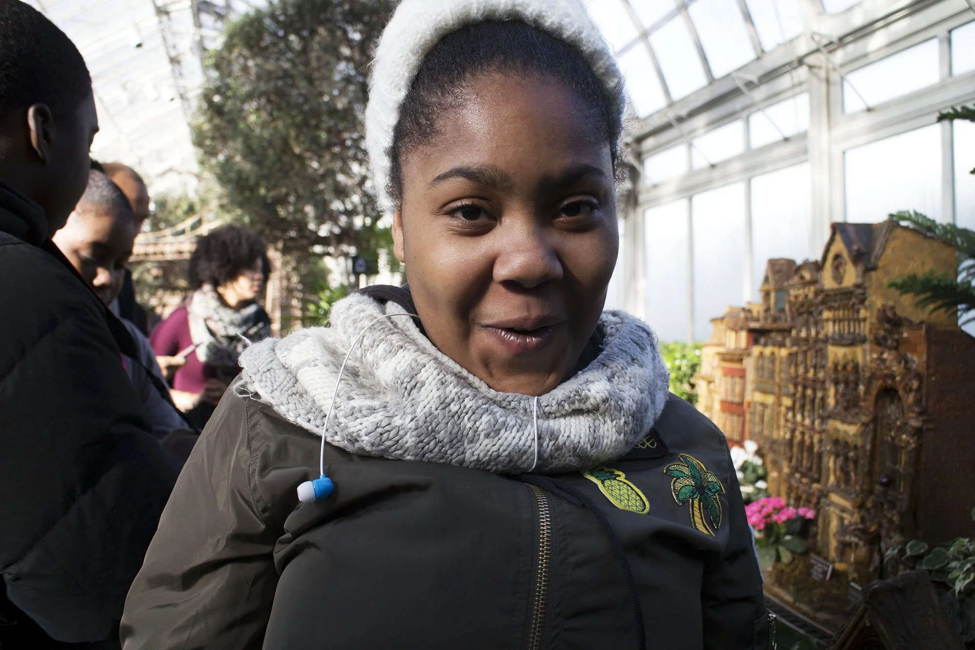 A woman with brown skin, wearing a grey knitted scarf and white fluffy hat, standing inside a greenhouse with a model of a building and pink flowers behind her.