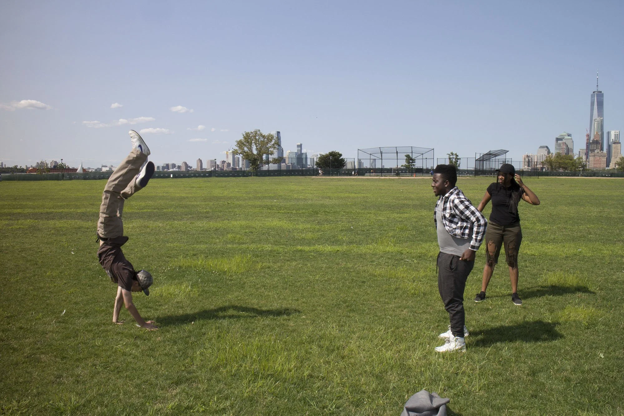 Three young people in a park. One is doing a handstand, another is smiling with hands on hips, and a woman stands behind, touching her hair. City skyline in the background.