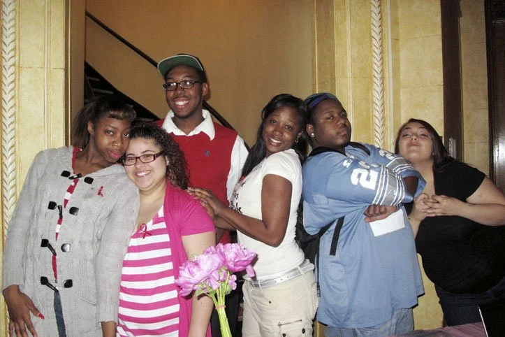 Group of six diverse young people standing together indoors, smiling and posing for a photo. One person is holding pink flowers.