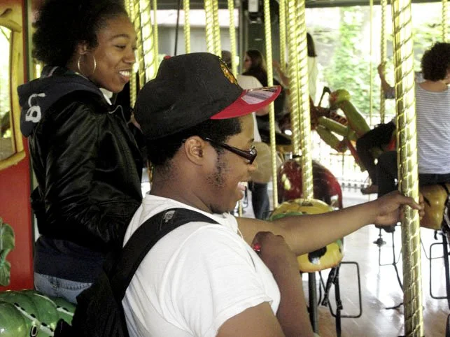 A young woman and a young man enjoying a ride on an amusement park carousel, with the young woman smiling and the young man sitting on a carousel horse, wearing a baseball cap and glasses.