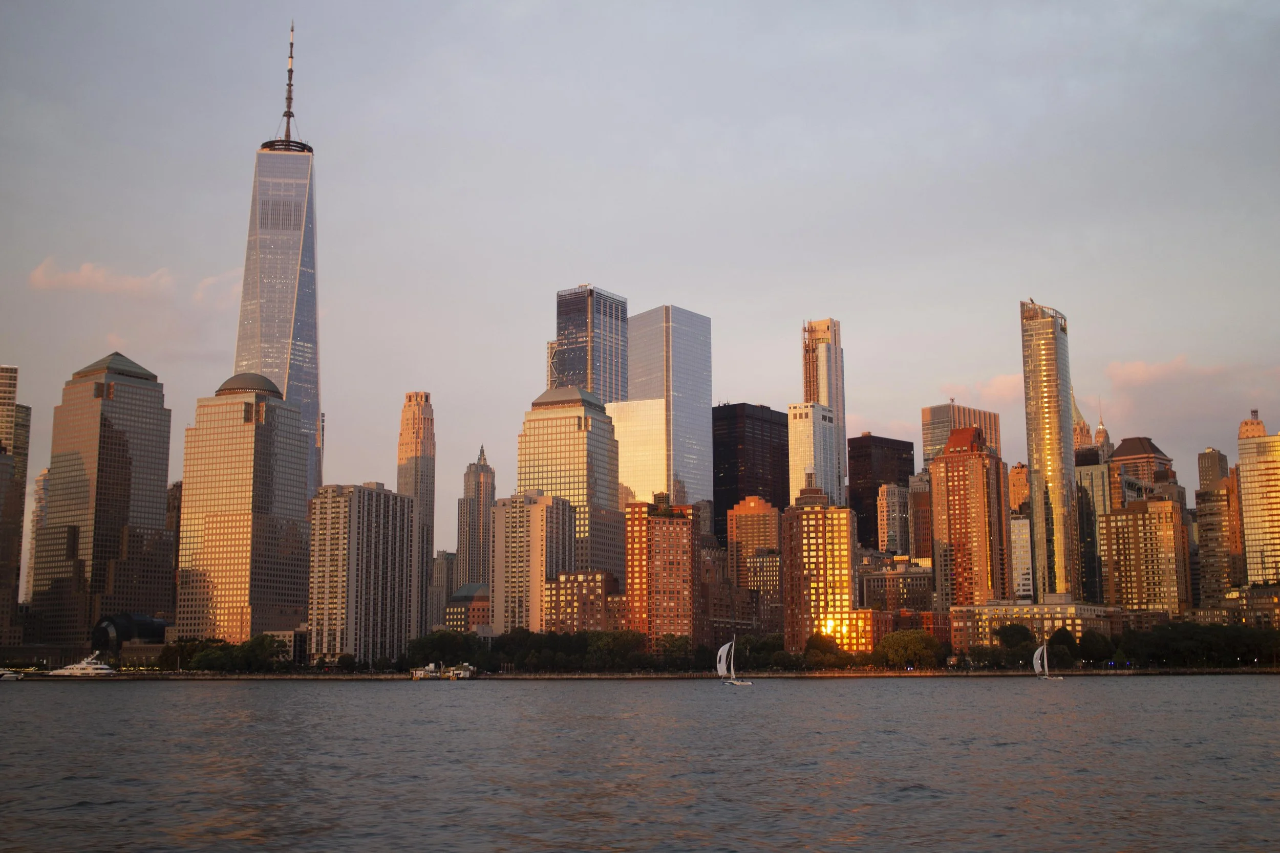 City skyline at sunset with tall skyscrapers, including One World Trade Center, view from across the water with sailboats