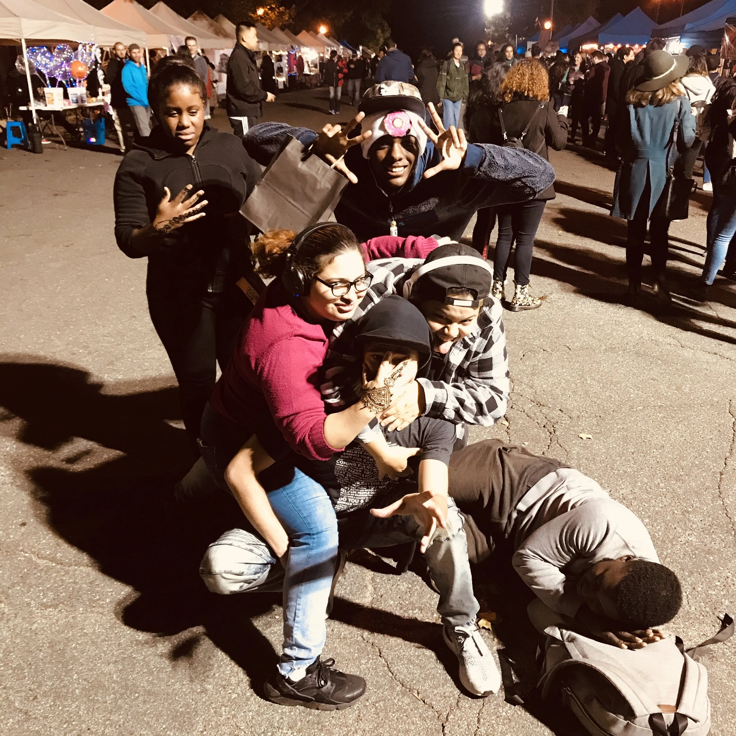 Group of six friends posing playfully at an outdoor night market with tents and people in the background.
