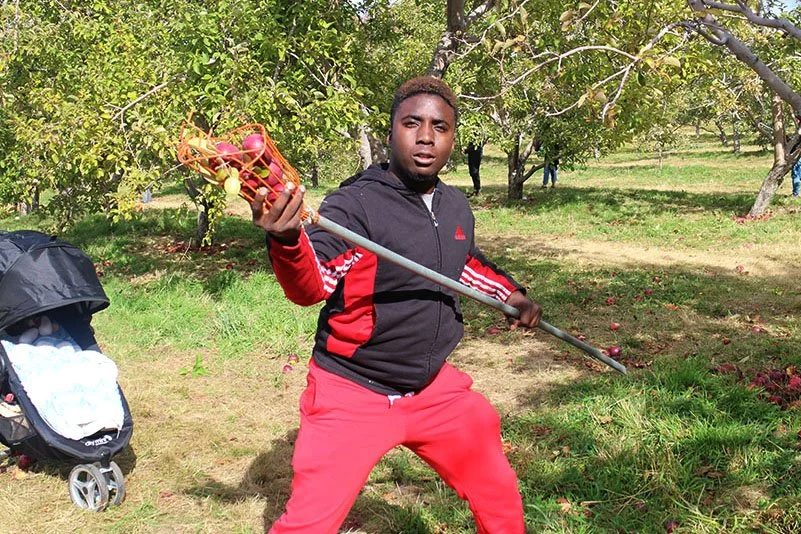 A young man in red and black outdoors holding a fruit picker stick with apples at an orchard.