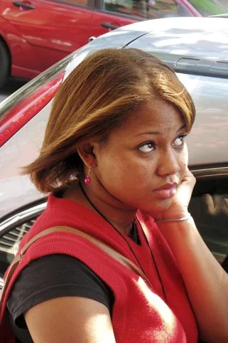 A young woman with shoulder-length brown hair, wearing a red vest over a black shirt, sitting next to a silver car and looking upwards with a thoughtful expression.