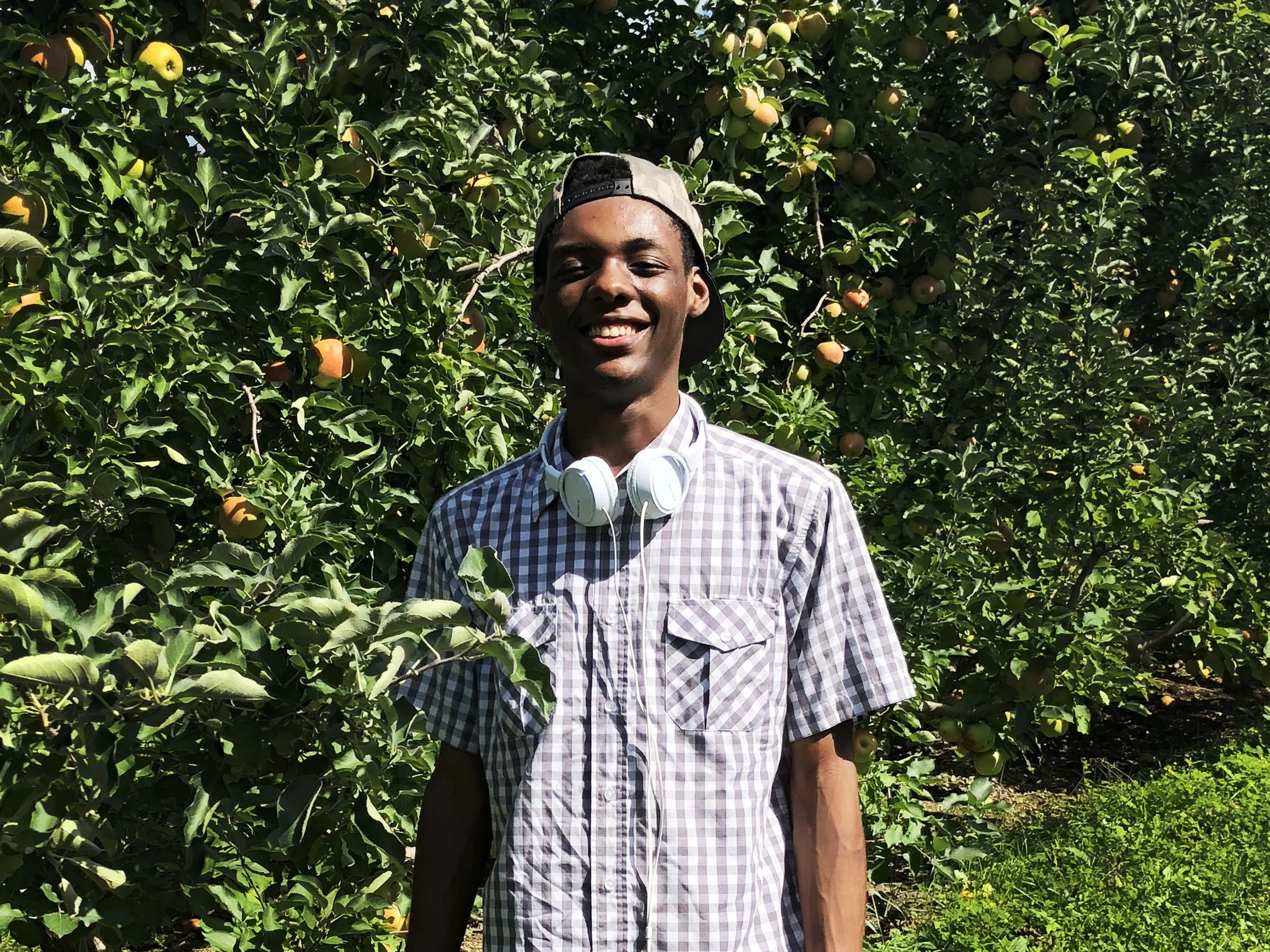 A young man standing in an apple orchard with a big smile, wearing a checked shirt, a backwards cap, and white headphones around his neck.