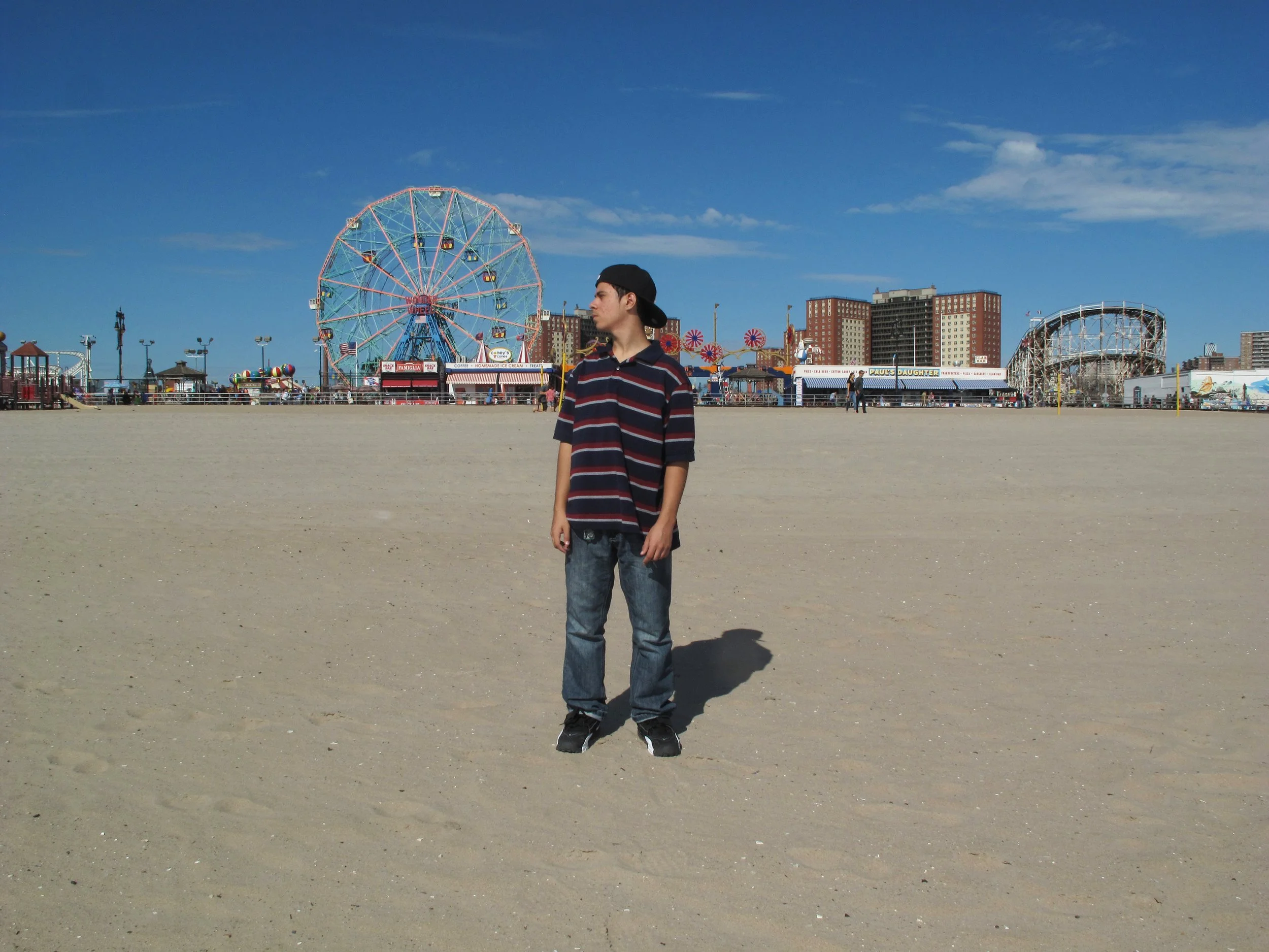 Young man in striped shirt and jeans standing alone on an empty beach, with a crowded amusement park and Ferris wheel in the background under blue sky.