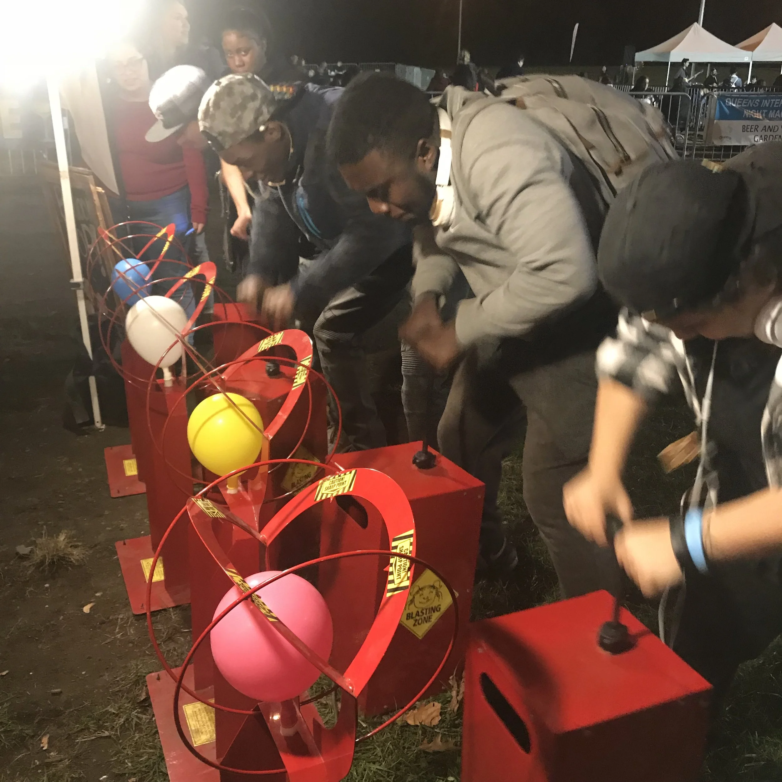 People playing a balloon popping game at a night event, with balloons inside red cages, some with warning labels, and a crowd in the background.