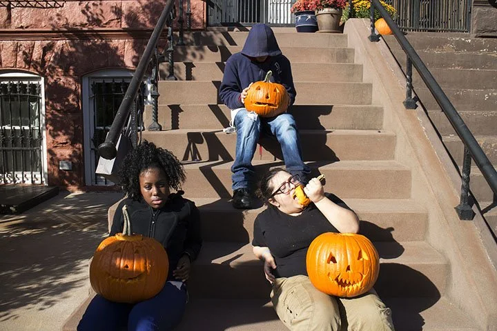 Three children sitting on steps with pumpkins. The child at the top holding a carved pumpkin, a girl with curly hair holding an uncarved pumpkin, and a girl with glasses eating a snack with a carved pumpkin on her lap.