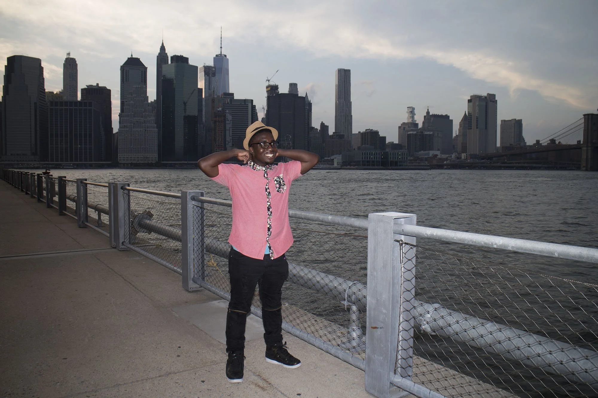 A person smiling and wearing glasses, a tan hat, a pink shirt with animal print accents, black pants, and sneakers, standing on a waterfront promenade with a city skyline in the background.