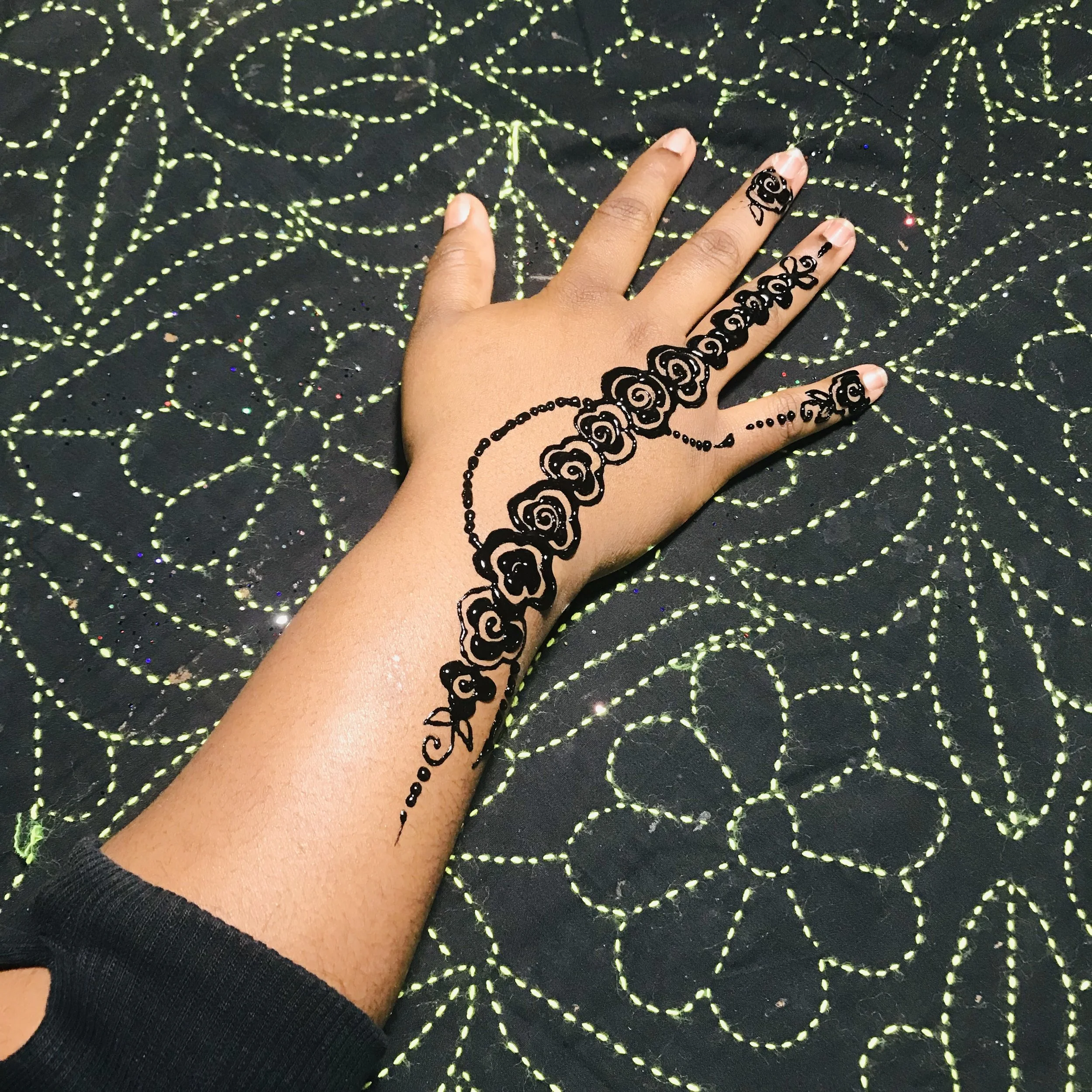 Hand with a black henna tattoo of roses and swirls on the back of the hand and wrist, resting on a dark fabric with neon green floral stitching.