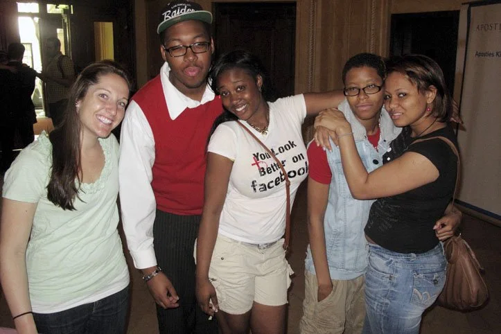 Group of five young people standing together indoors, smiling and hugging.