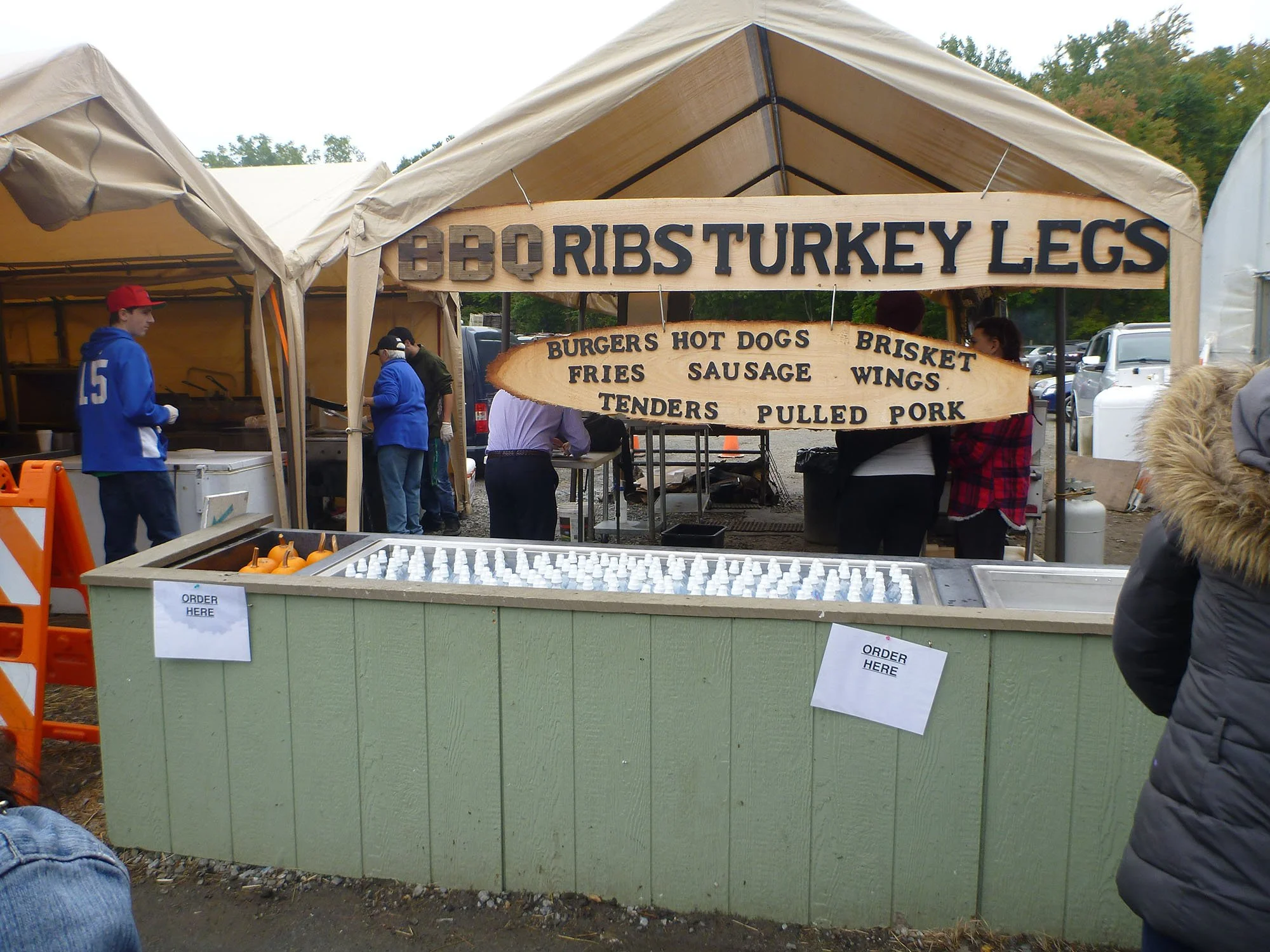 Food stand at outdoor fair with a wooden sign listing BBQ ribs, turkey legs, burgers, hot dogs, fries, sausage, brisket, wings, tenders, and pulled pork, serving food to customers.