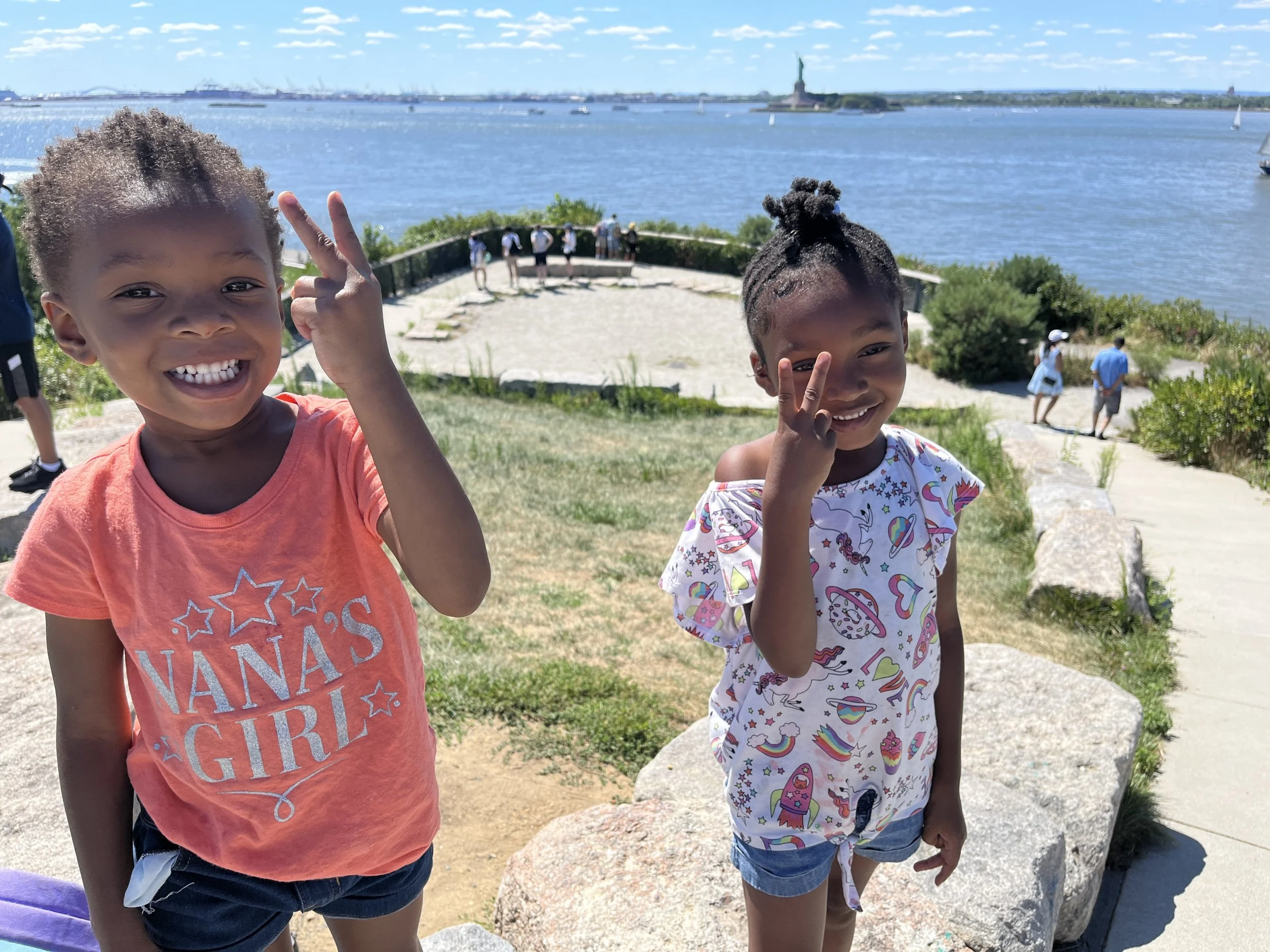 Two young girls smiling and making peace signs with their hands outdoors near the water, with the Statue of Liberty visible in the background.