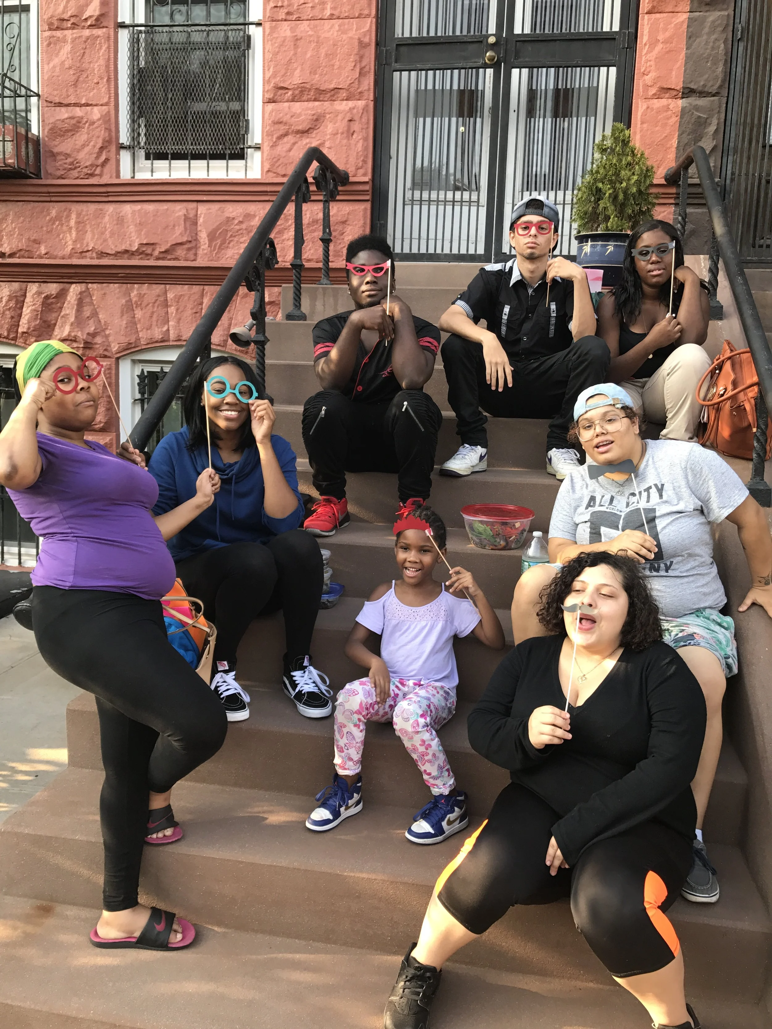 Group of diverse young people sitting on steps outside, some holding paper mustaches and glasses on sticks for fun, with a rental bracelet and casual clothing.