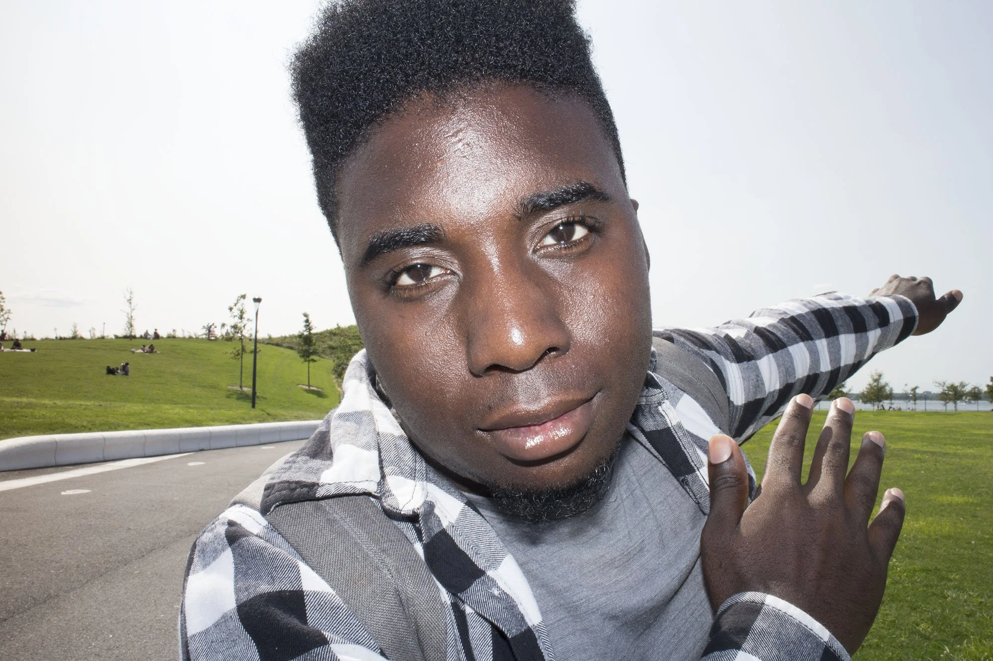 Young man with dark skin and short curly hair pointing to the right in an outdoor park setting.
