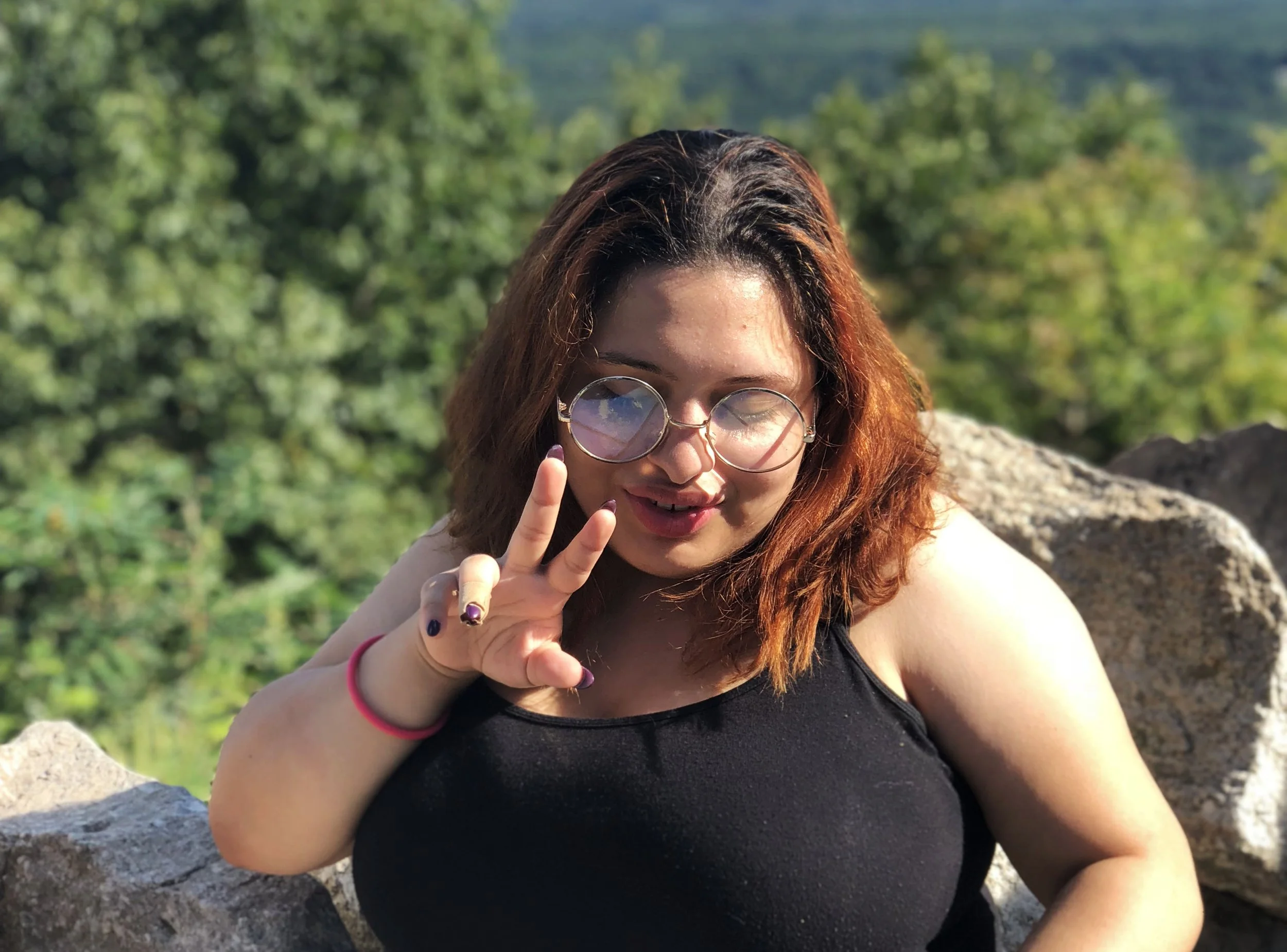 A young woman with reddish-brown hair, wearing round glasses and a black sleeveless top, making a peace sign with her right hand. She is outdoors with a background of green trees and rocky terrain.
