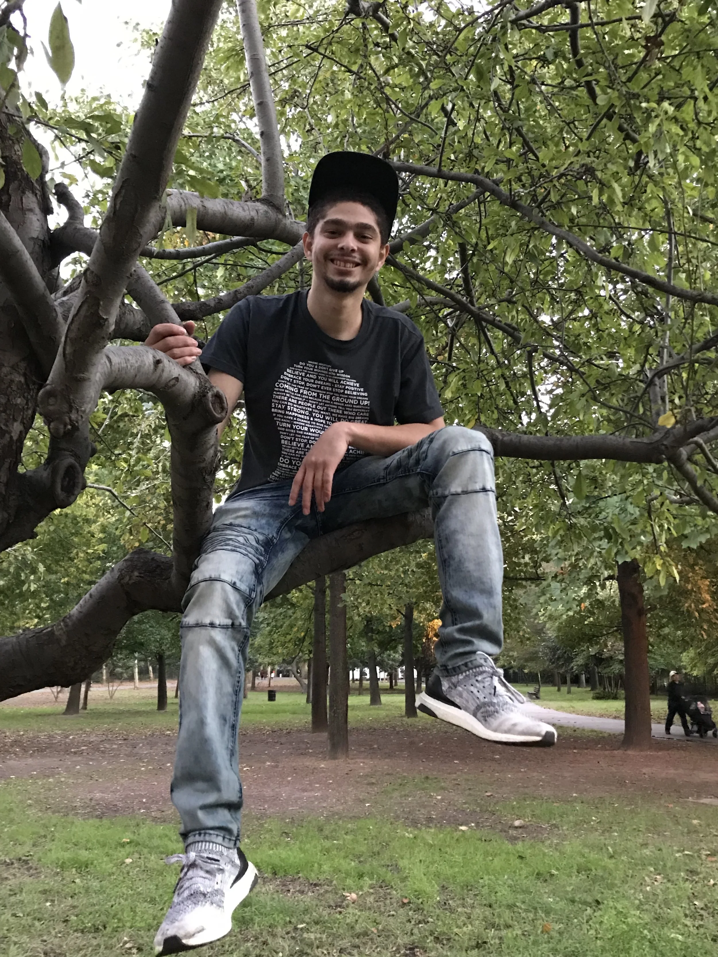 A young man with dark hair and a beard, wearing a black T-shirt, jeans, and sneakers, is sitting on a tree branch in a park surrounded by green trees and grass, smiling at the camera.