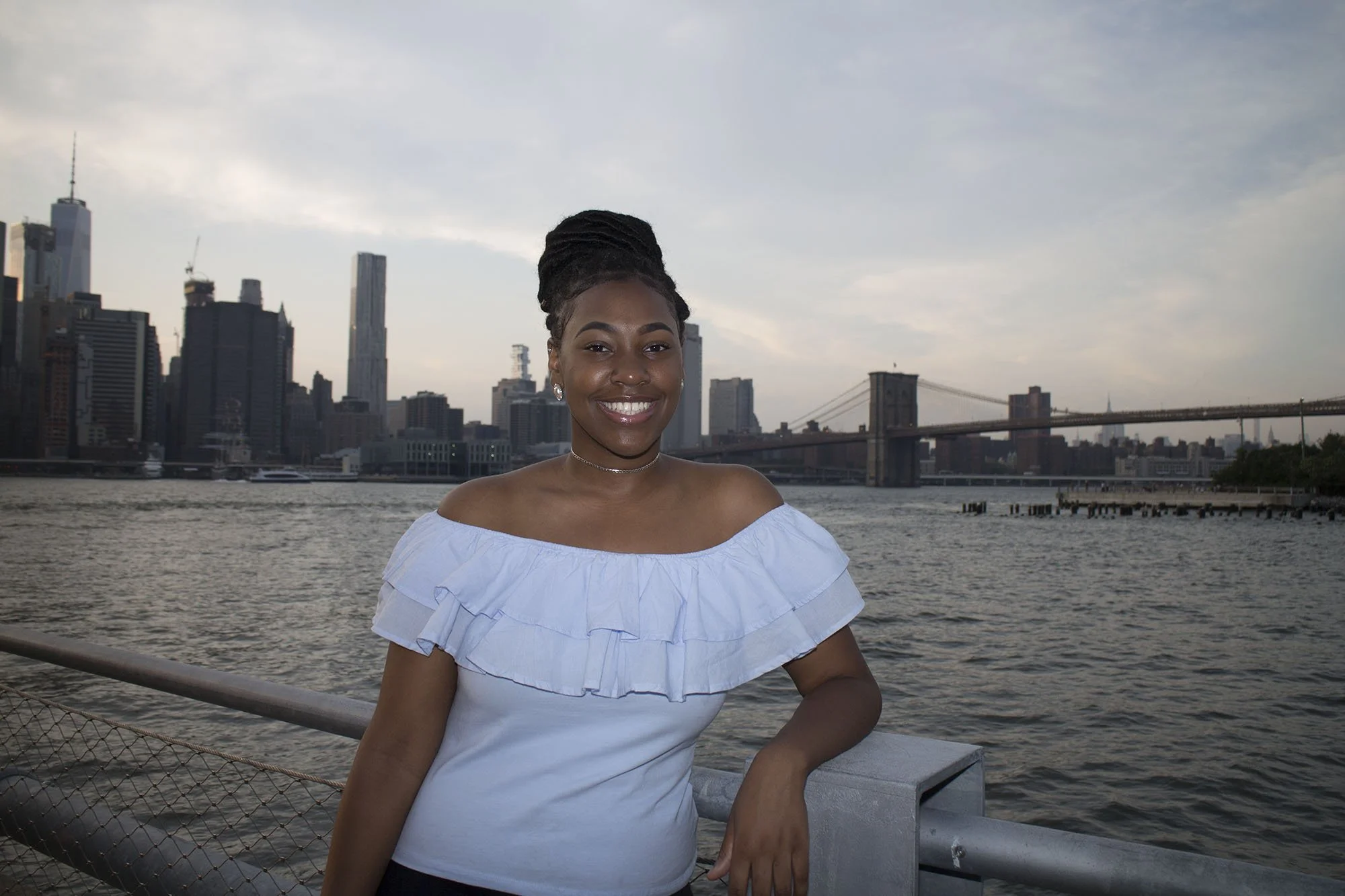A smiling woman with dark hair styled in an updo standing by the river in front of a city skyline and a bridge, wearing an off-the-shoulder white top.