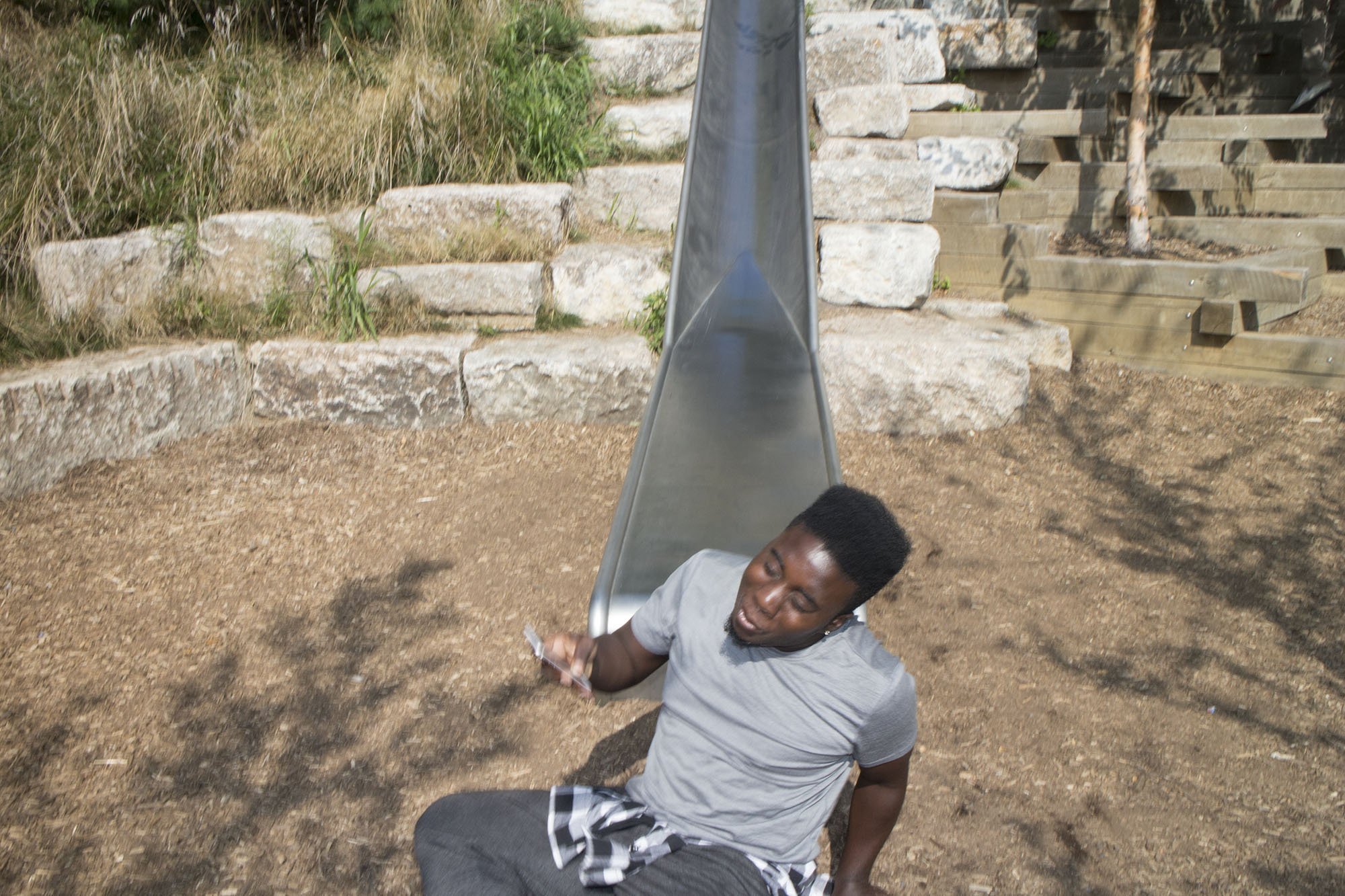 A young man sitting on the ground at the base of a metal slide at a playground, holding a phone and looking down, with a gravel surface and stone steps in the background.