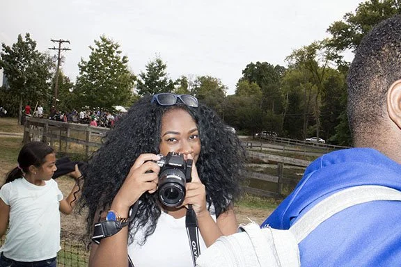 Young woman with curly hair taking a photo with a camera at an outdoor event.