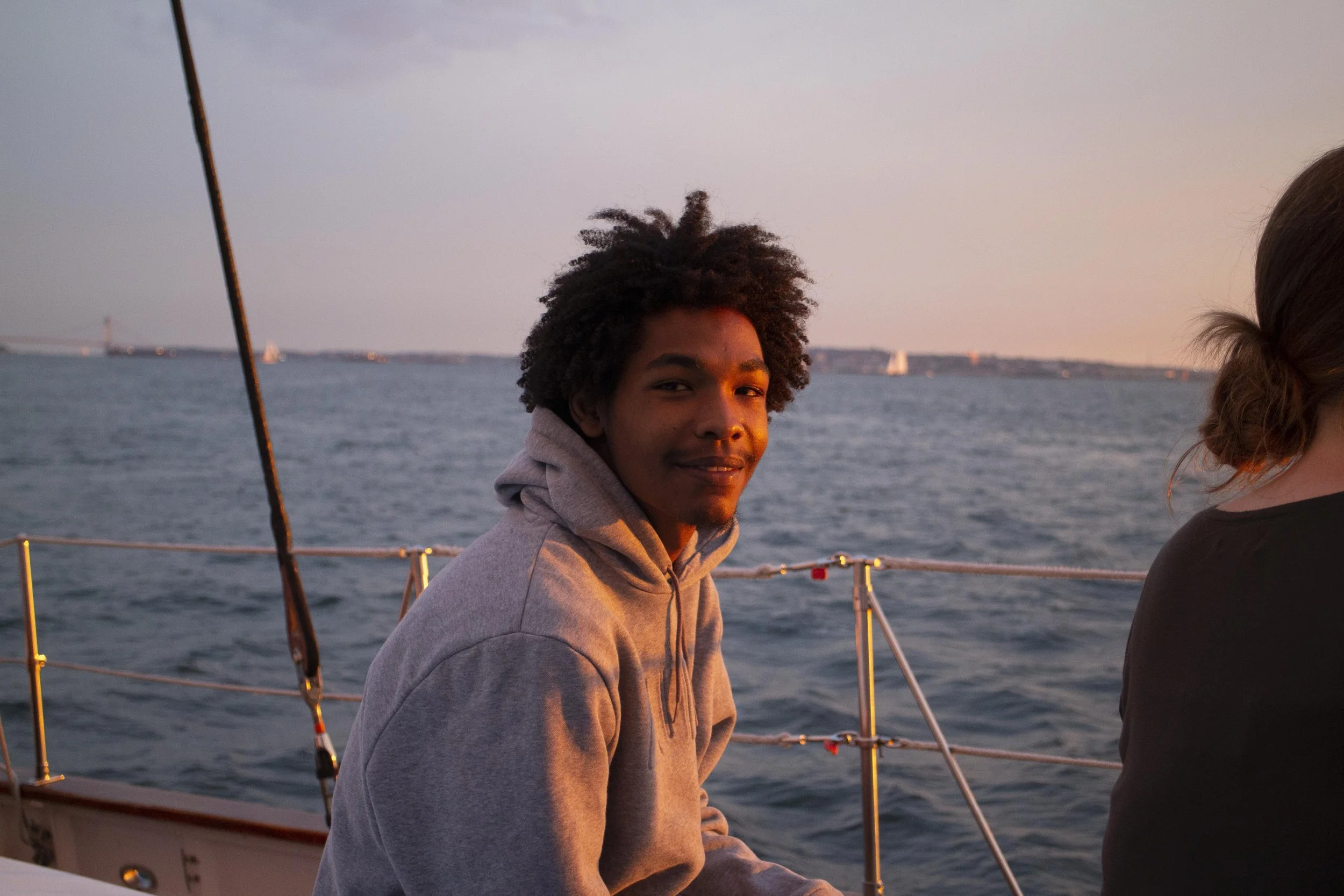 A young man with curly hair wearing a gray hoodie sits on a boat during sunset, smiling at the camera with the ocean and distant land in the background.