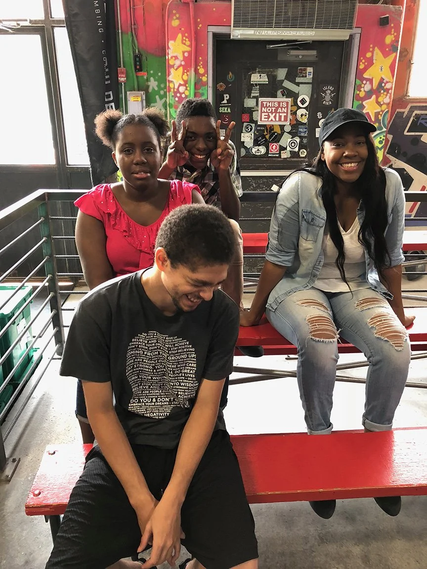 Four young people, two boys and two girls, are sitting and standing on a red picnic table inside a colorful room with graffiti-style wall art. They are smiling and making playful gestures.