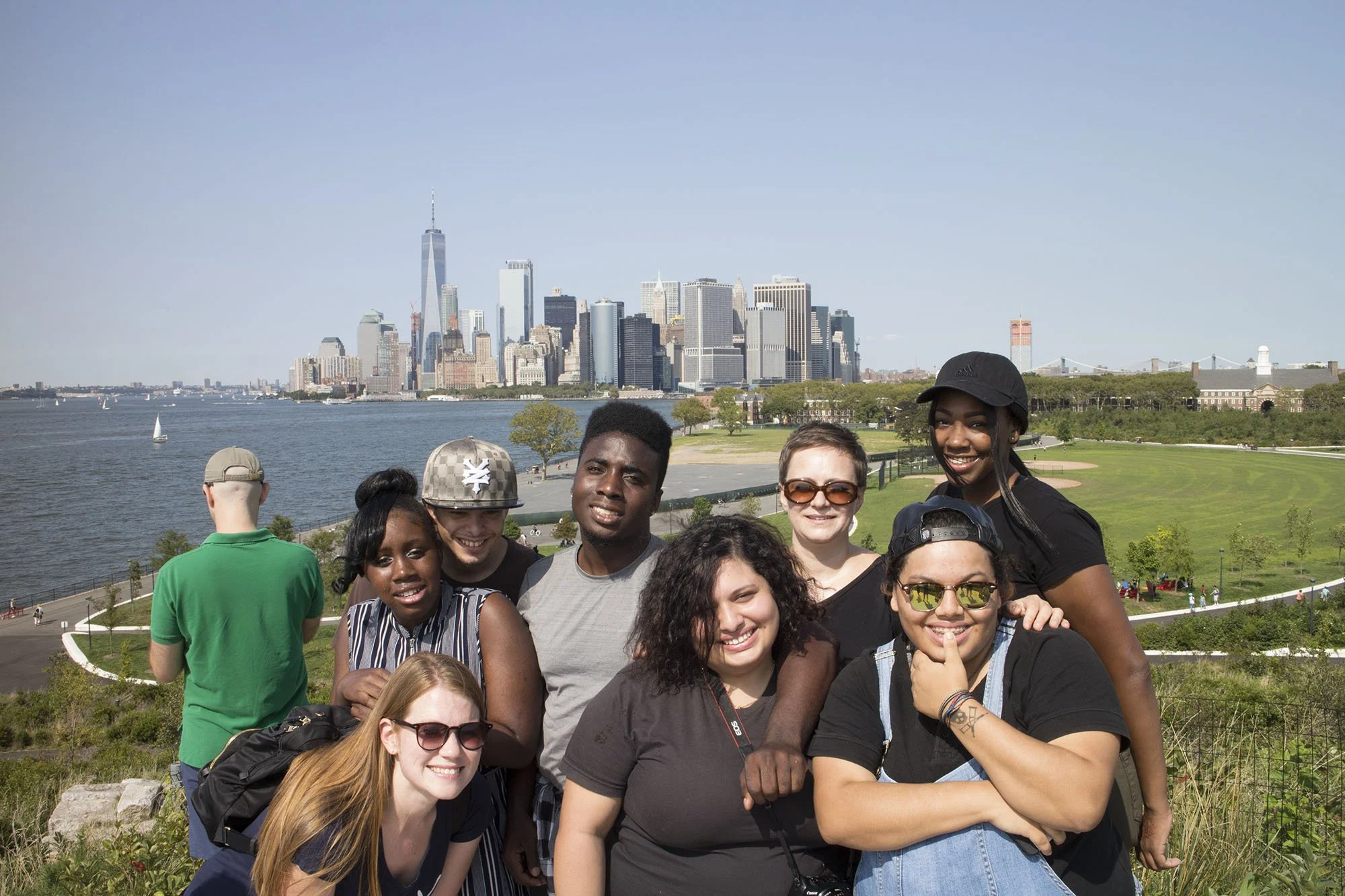 A diverse group of people smiling and posing for a photo outdoors near a city skyline, with a body of water and grass area in the background.
