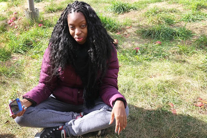 Young woman sitting cross-legged outdoors on a grassy area, holding a smartphone, with a relaxed pose and casual clothing.