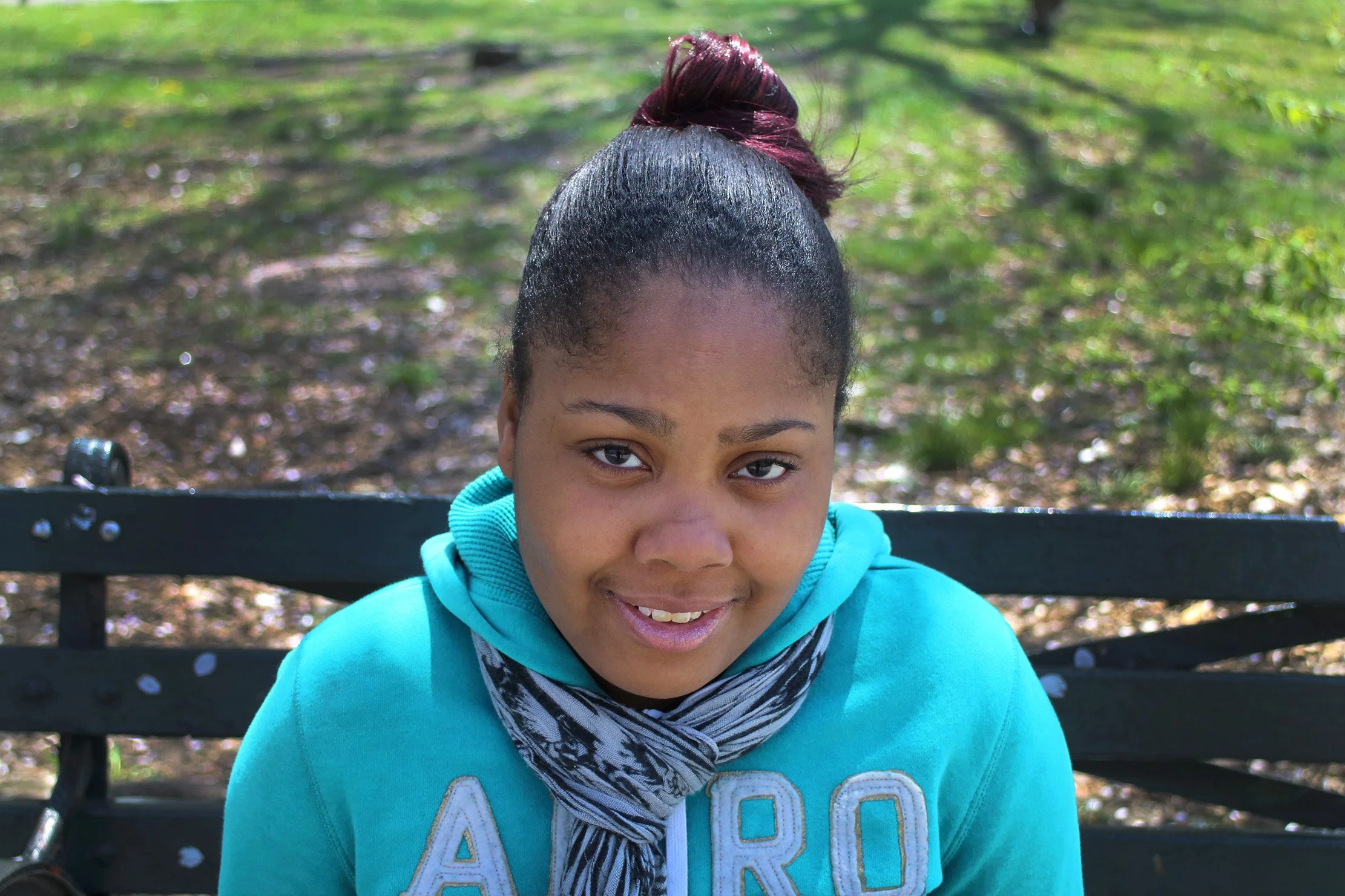 A young woman with dark hair in a bun, wearing a blue hoodie and a scarf, sitting on a black park bench with a background of green trees.