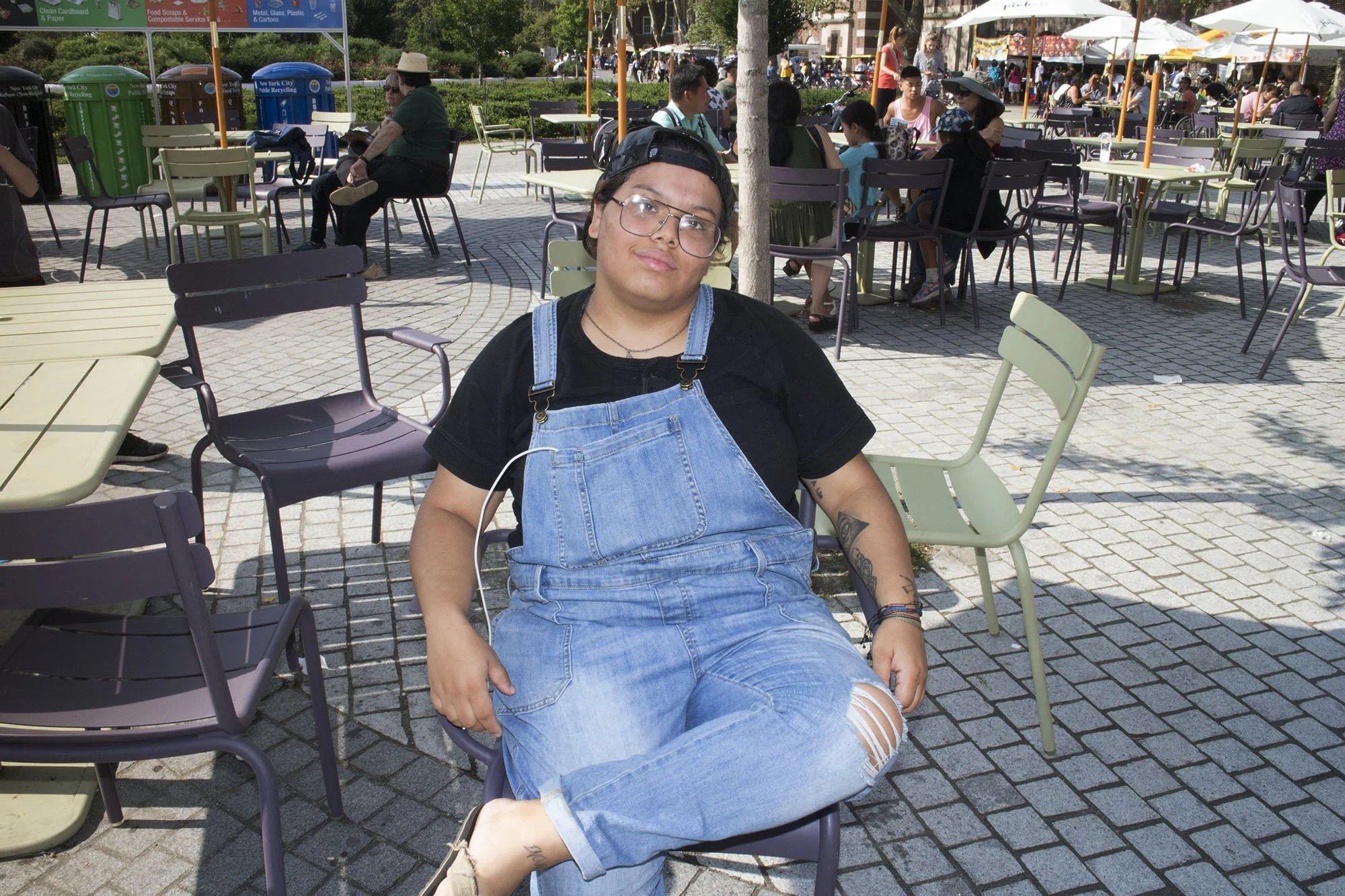 Person sitting on a chair outdoors at a cafe in a city square, with tables, chairs, umbrellas, and other people in the background.