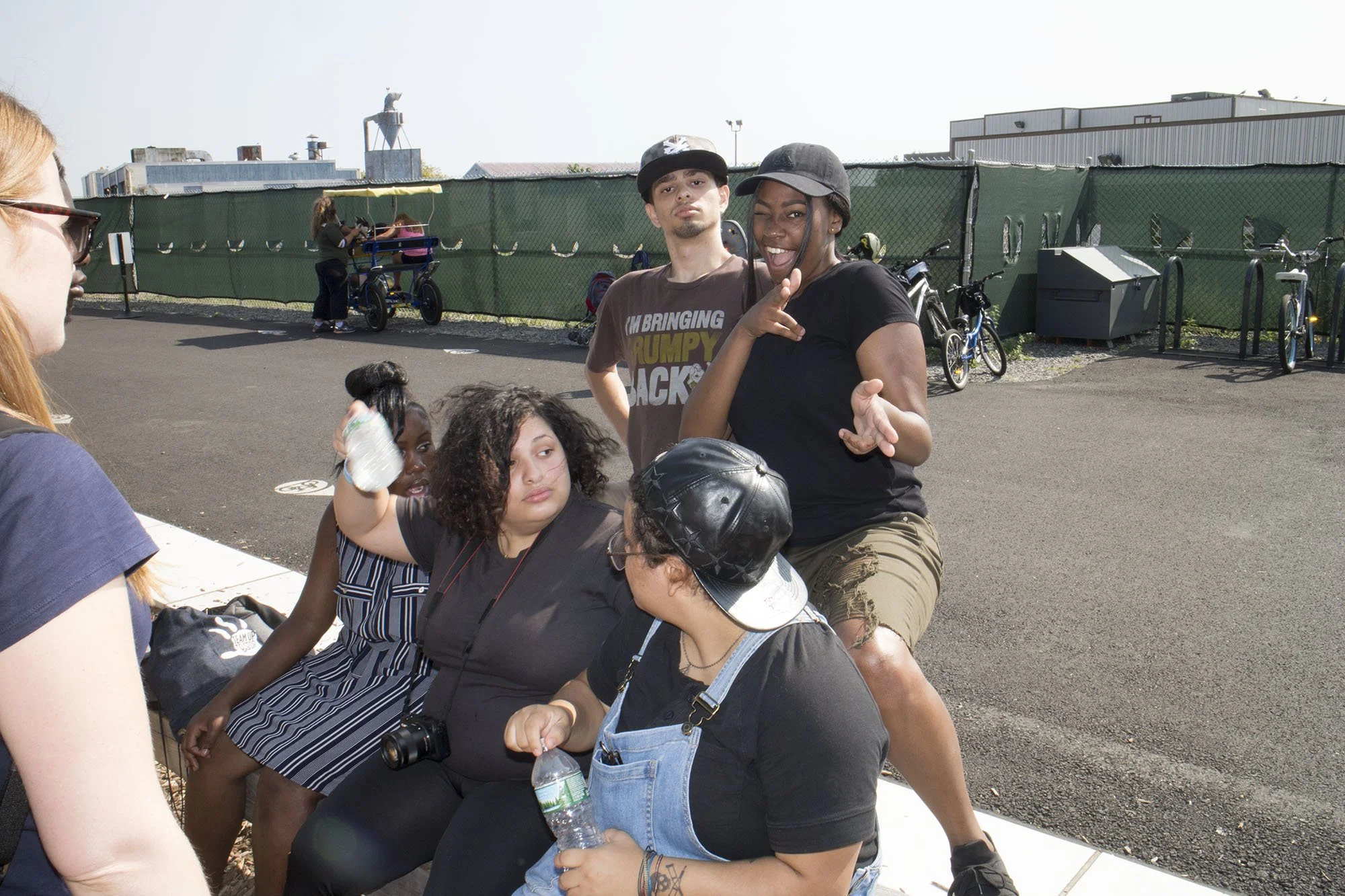 Group of young people sitting and standing outdoors near a parking lot with bicycles and a green fence in the background. One person is pointing and another is making a finger gun gesture.