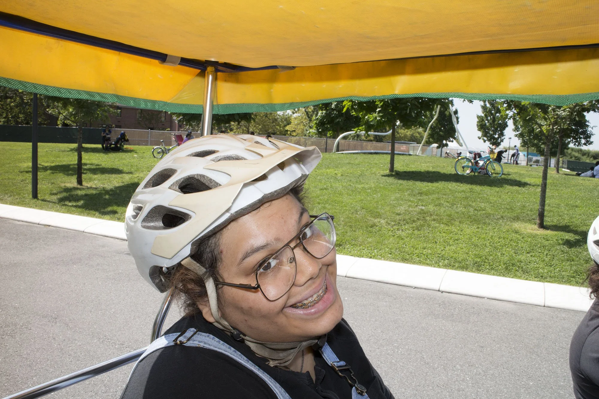 Young person smiling, wearing glasses and a bike helmet, riding a pedal-powered vehicle with a yellow canopy at a park.