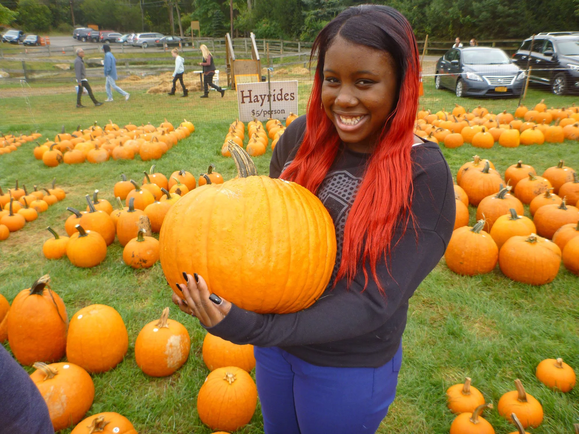 Woman with long red hair smiling while holding a large orange pumpkin at a pumpkin patch with many pumpkins on the grass. There are cars and people in the background.