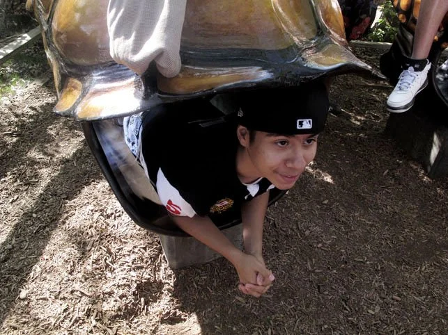 Child with a baseball cap crouched down in a small pipe or tunnel at an outdoor park or playground.