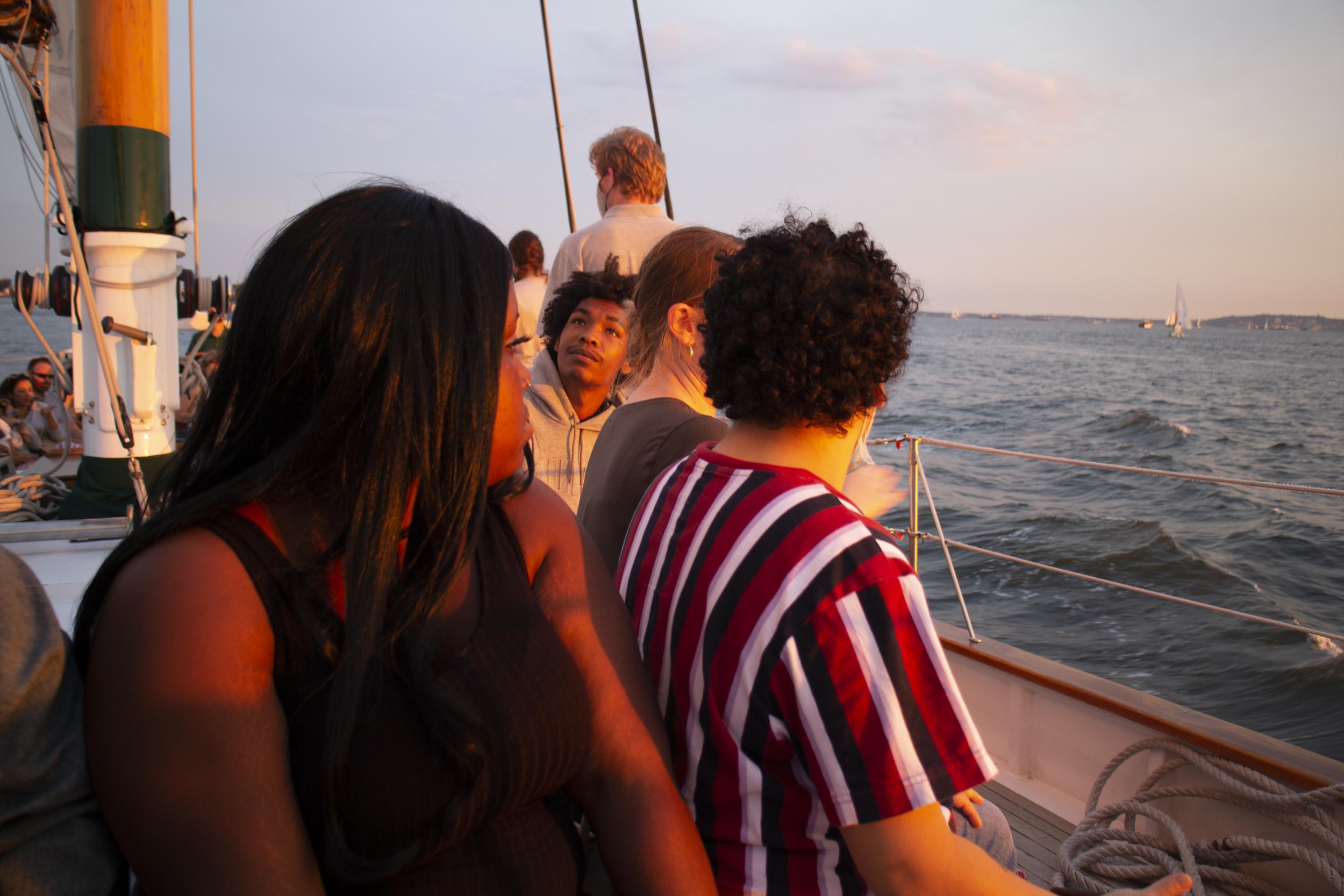 Group of people on a sailboat during sunset looking out at the water with sailboats in the distance.