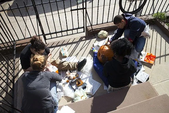 Four people sitting on a sidewalk, carving a large pumpkin with various tools and supplies around them, enclosed by a metal fence.