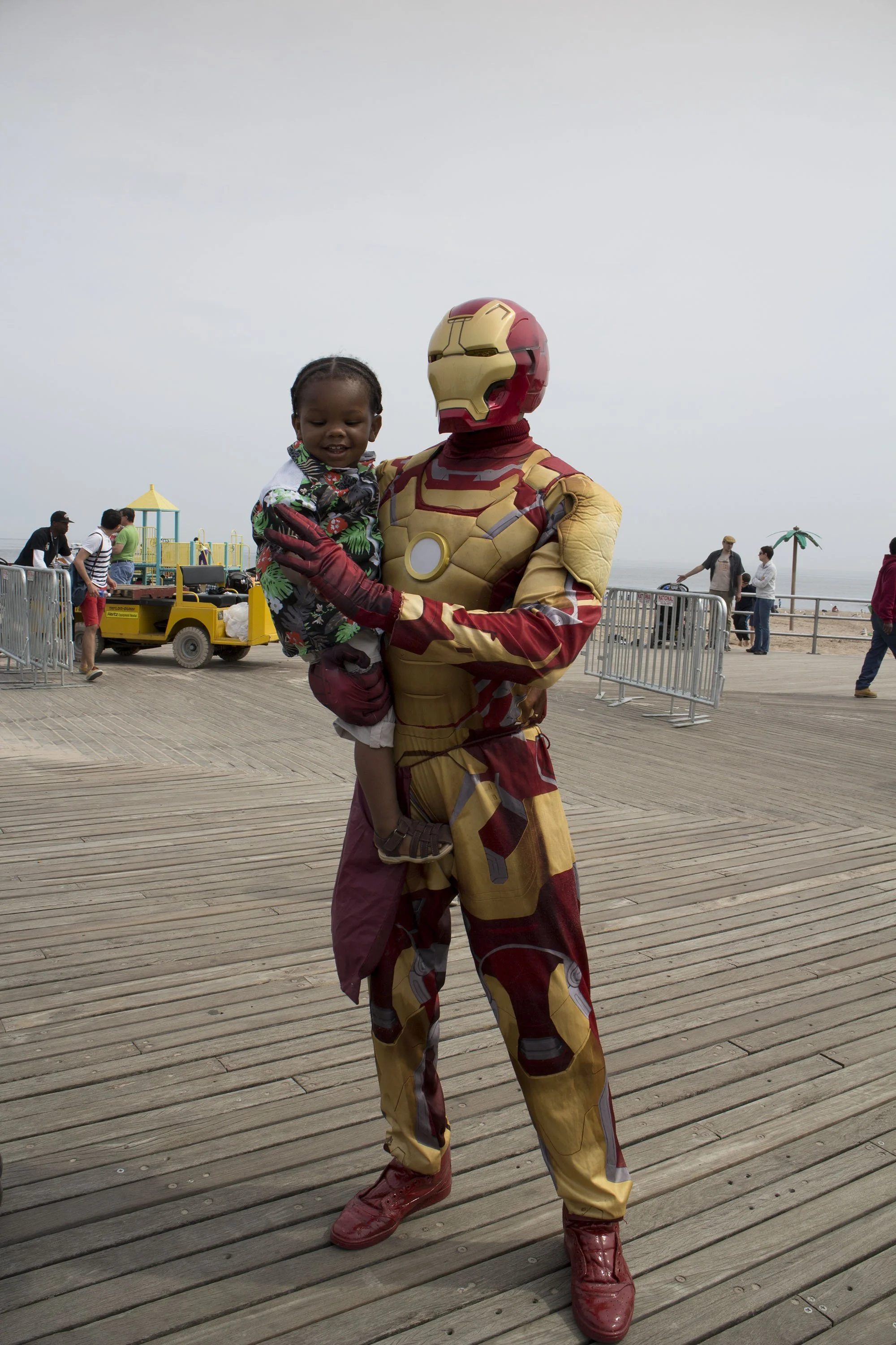 Person dressed as Iron Man holding a young child on a pier, with a beach and overcast sky in the background.