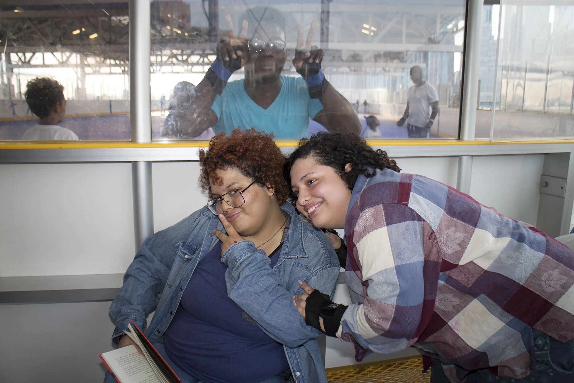 Two young women sitting close together at an outdoor rink, one with glasses and curly hair holding a book, the other smiling and leaning in, with a man making peace signs behind a glass barrier in the background.