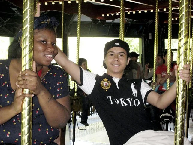 Two people sitting on a carousel ride, smiling at the camera, with other park visitors in the background.