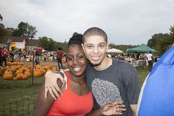 A smiling young woman and man hugging at an outdoor pumpkin festival, with pumpkins and tents in the background.