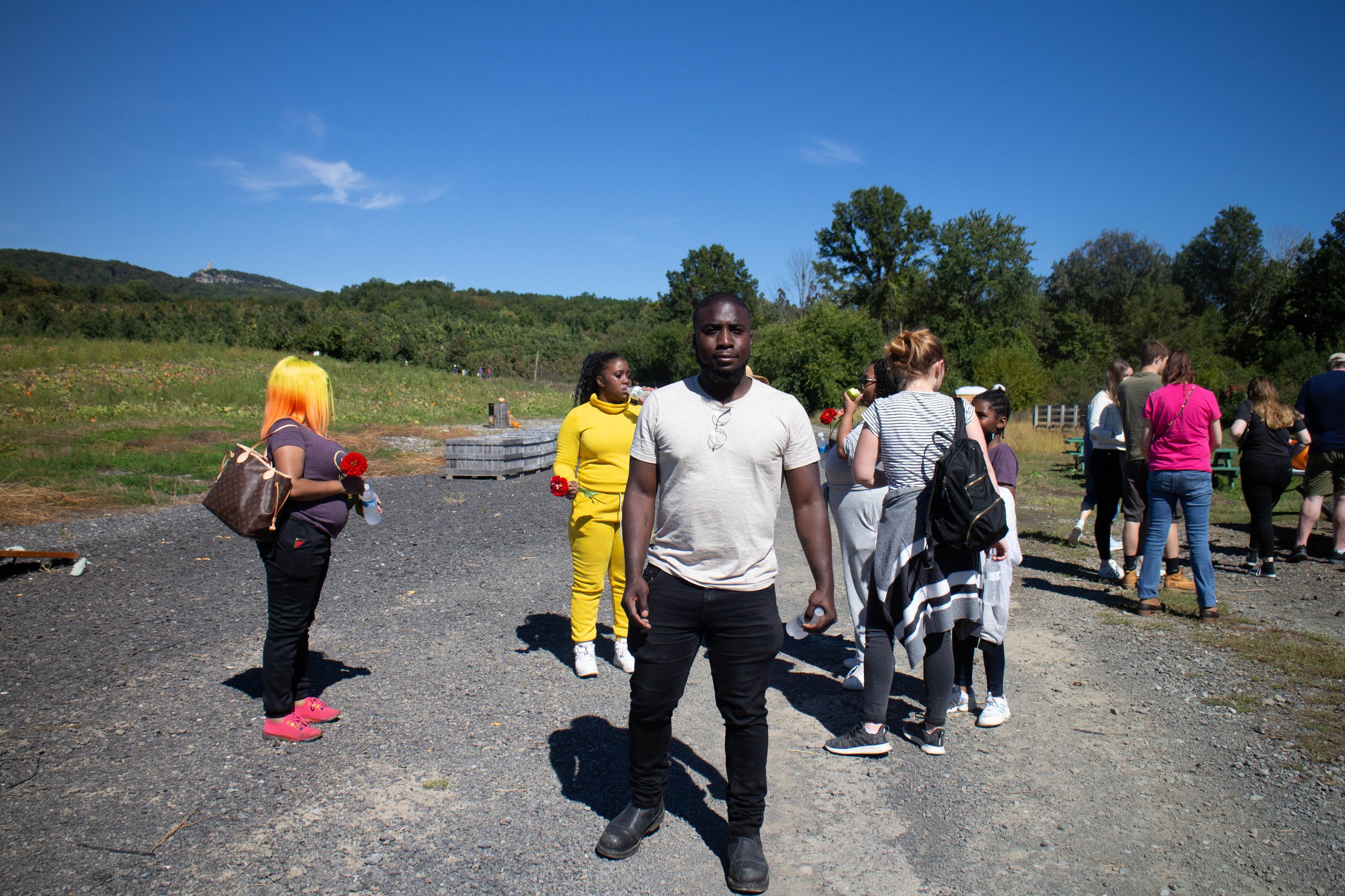 Group of diverse people gathered outdoors on a sunny day, some holding flowers, with trees and a hill in the background.
