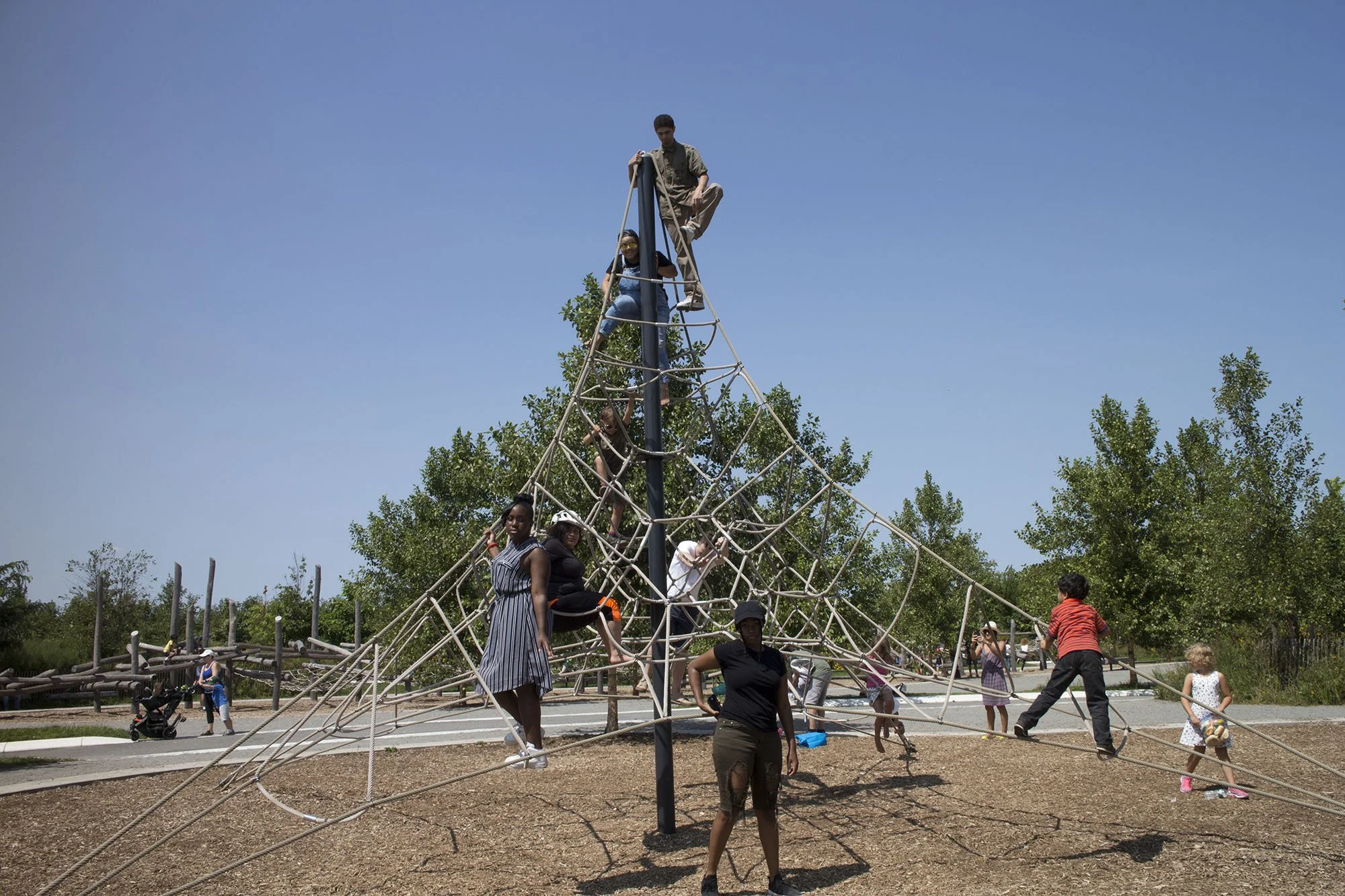 Children and adults playing on a large rope climbing structure in a park, with trees and a paved pathway in the background, under a clear blue sky.