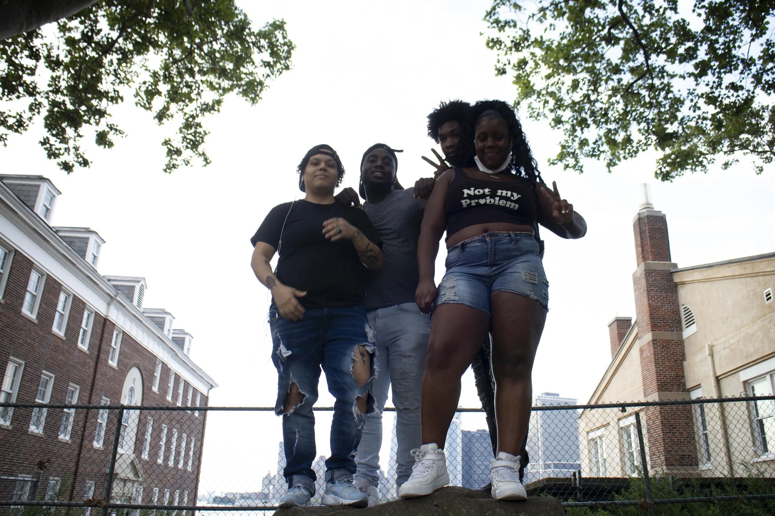 Four young people standing together outdoors on a fence, with buildings and trees in the background, smiling and making peace signs.