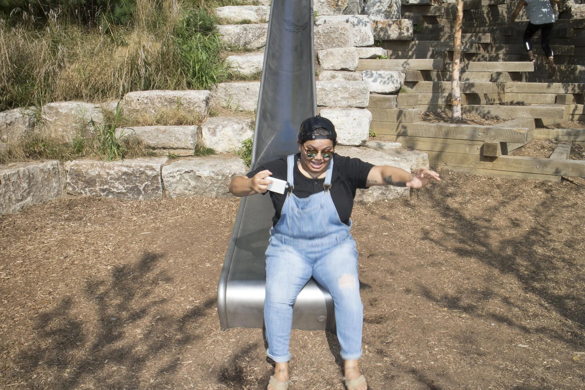 Woman sitting on a playground slide, holding a phone, smiling, and reaching out with her left hand, outdoors with dirt ground, stone steps, and wooden stairs in the background.