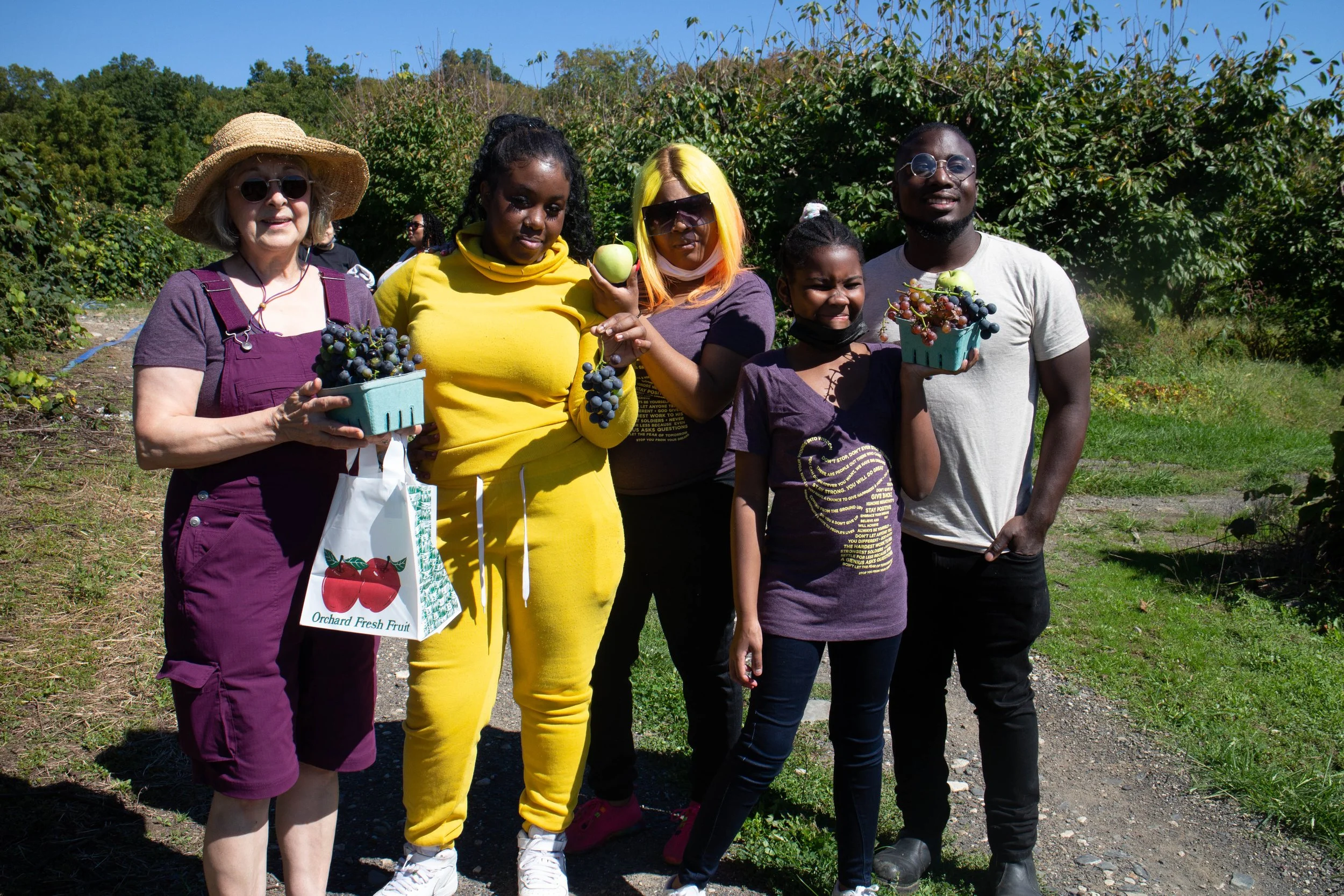 Group of five people standing in a vineyard holding baskets of grapes and fruit, smiling on a sunny day.