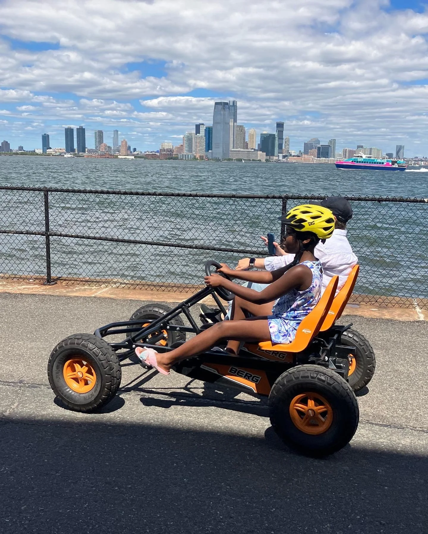 A young girl in a yellow bike helmet and a sleeveless dress riding a quad bike along a waterfront, with a woman behind her, possibly her mother, taking a picture, and a city skyline with tall buildings and a boat on the water in the background.