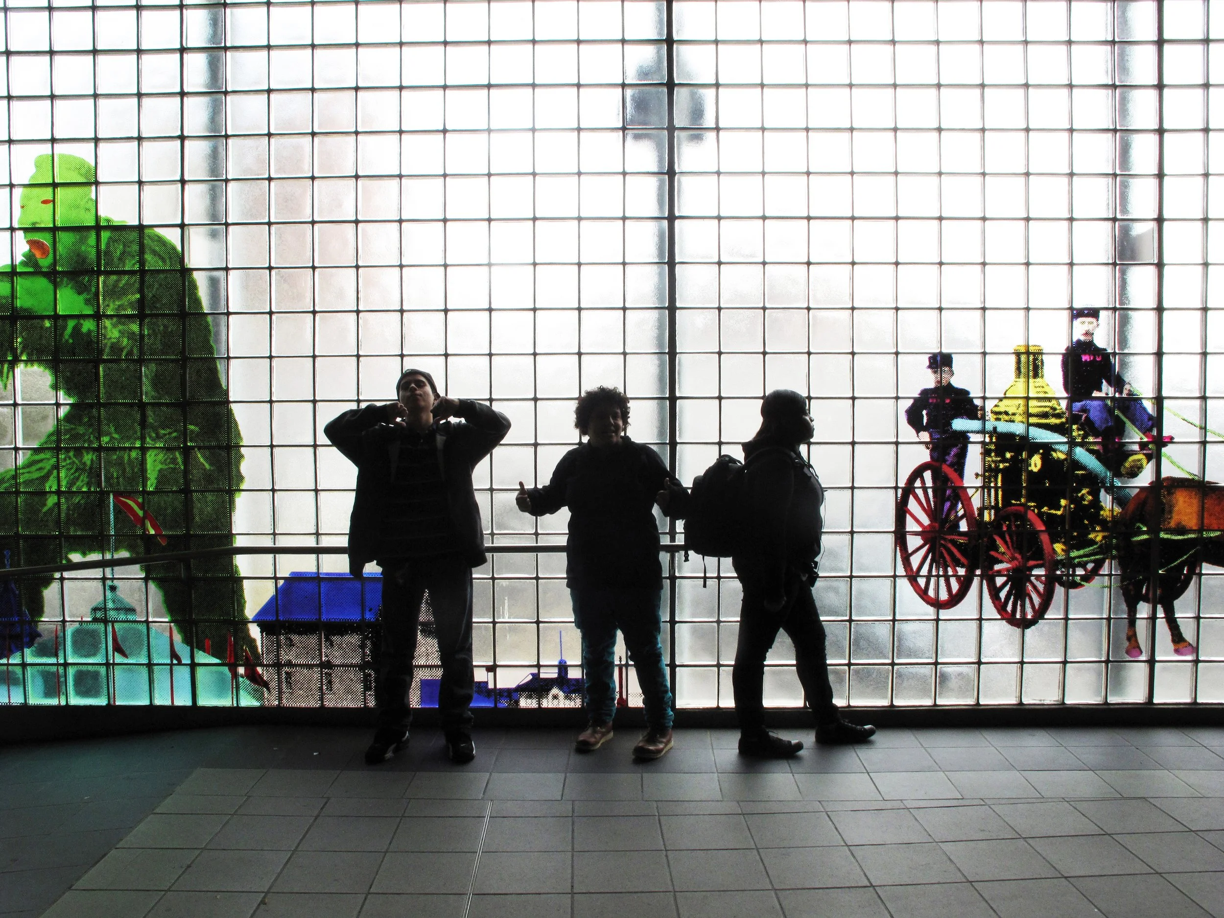 Silhouettes of three people standing in front of a large glass window with colorful decorations and figures visible outside, including a large green dinosaur and a horse-drawn carriage with two people riding inside.