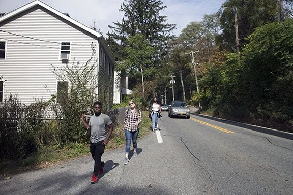 Three people walking along the side of a residential street with cars and trees, with a house on the left and utility poles on the right.