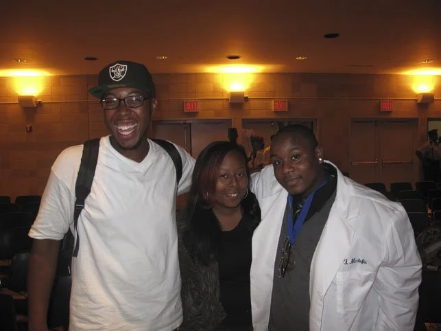 Three people smiling and posing together indoors at an event, with chairs visible in the background.